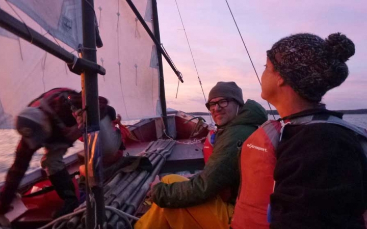 Three people wear life jackets in a sailboat under a pink and purple sky.